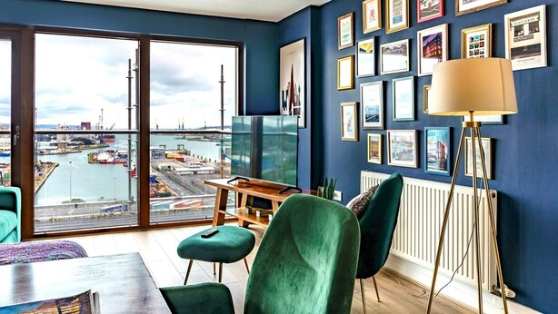 A modern room with a large window overlooking a port, a dining area with green chairs, and a wall decorated with framed pictures. - City Centre | Top Floor w/ Balcony & Sunset Views | Pass the Keys (Belfast)