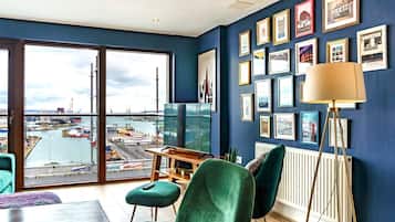 Une chambre moderne dotée d’une grande fenêtre donnant sur un port, d’un coin repas avec des chaises vertes et d’un mur décoré de tableaux encadrés.
