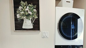 Un área de lavandería con dos lavadoras apiladas, un carro azul turquesa con productos de limpieza y un cuadro con flores en la pared.