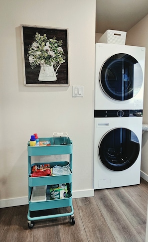 Un espace buanderie comprenant deux machines à laver superposées, un chariot turquoise contenant des produits de nettoyage et un tableau floral encadré accroché au mur.