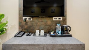 A reception area with a flat-screen TV mounted on a wall, a telephone, two mugs, a tray with a kettle, water bottles, and a plant. - Lime Tree Hotel Medanta (Gurugram)