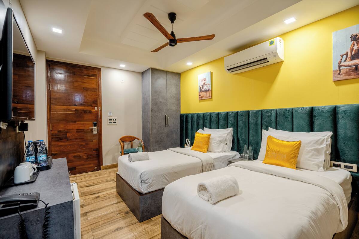 A hotel room with two beds, a desk with a telephone, a ceiling fan, an air conditioning unit, and a painting on the wall.