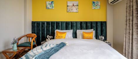 Una habitación de hotel con una cama grande, una silla de madera, una mesita pequeña y una obra de arte en la pared amarilla.