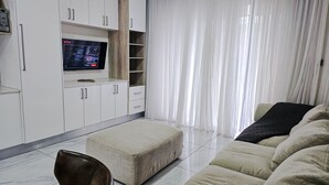 En moderne stue med et fladskærms-tv monteret på et hvidt skab, en beige sofa, en læderstol i brunt skind og et stort vindue med lette, hvide gardiner.