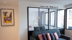 En moderne stue med en grå sofa, et indrammet kunstværk på væggen og et stort vindue med persienner.