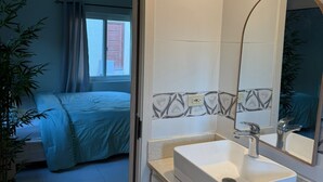 Un baño de hotel con un lavamanos blanco, un espejo y una puerta que da a un dormitorio con cama y cortinas.