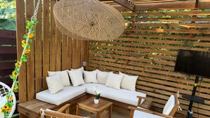 Una terraza de madera con un sofá seccional blanco, sillas de madera, una lámpara colgante de mimbre y un televisor montado en una pared de madera.