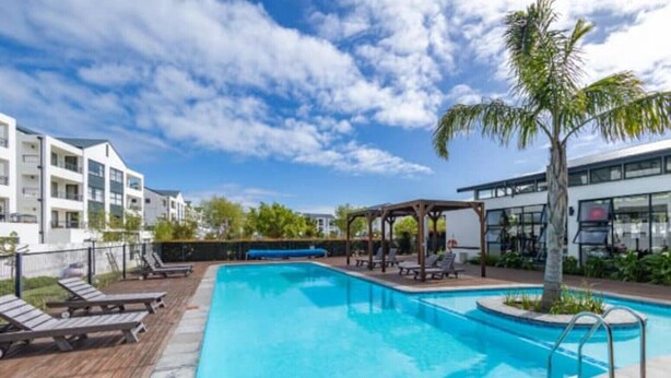 Una piscina rectangular con muelle de madera y sillones de descanso, rodeada de edificios modernos y un cielo despejado.