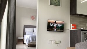 Un dormitorio moderno con cama, televisor mostrando 'TOP HITS 2024' y una pequeña zona de kitchenette.