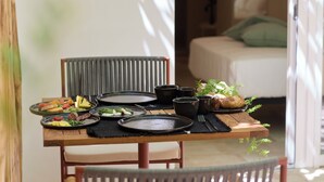A dining table set with plates, bowls, and utensils, with a bed and a window in the background. - Musae In Lama (Polignano a Mare)