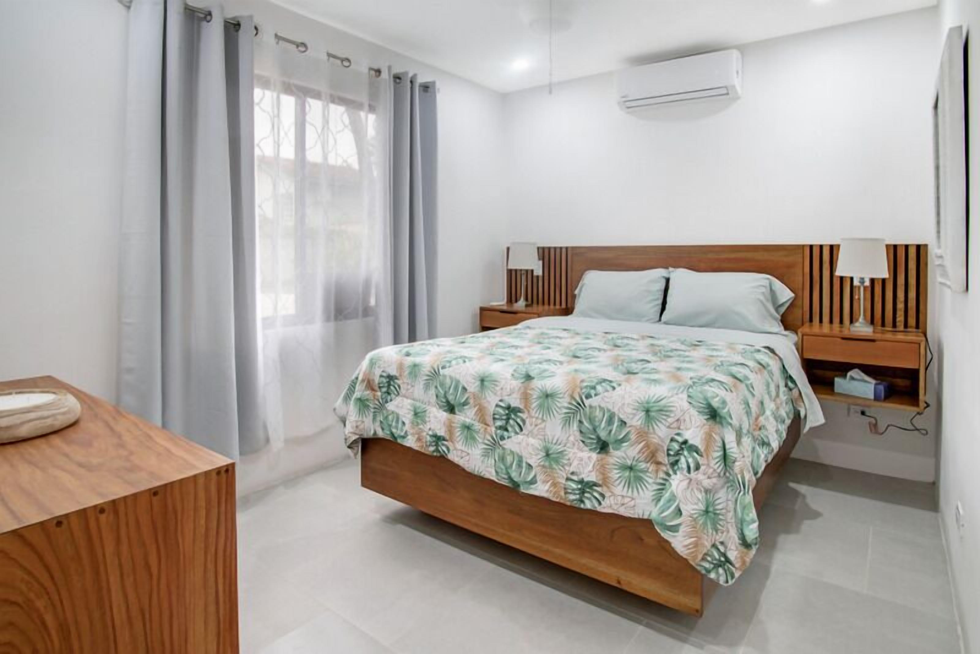Un dormitorio con cama, una mesita de noche, cortinas en la ventana y un equipo de aire acondicionado.