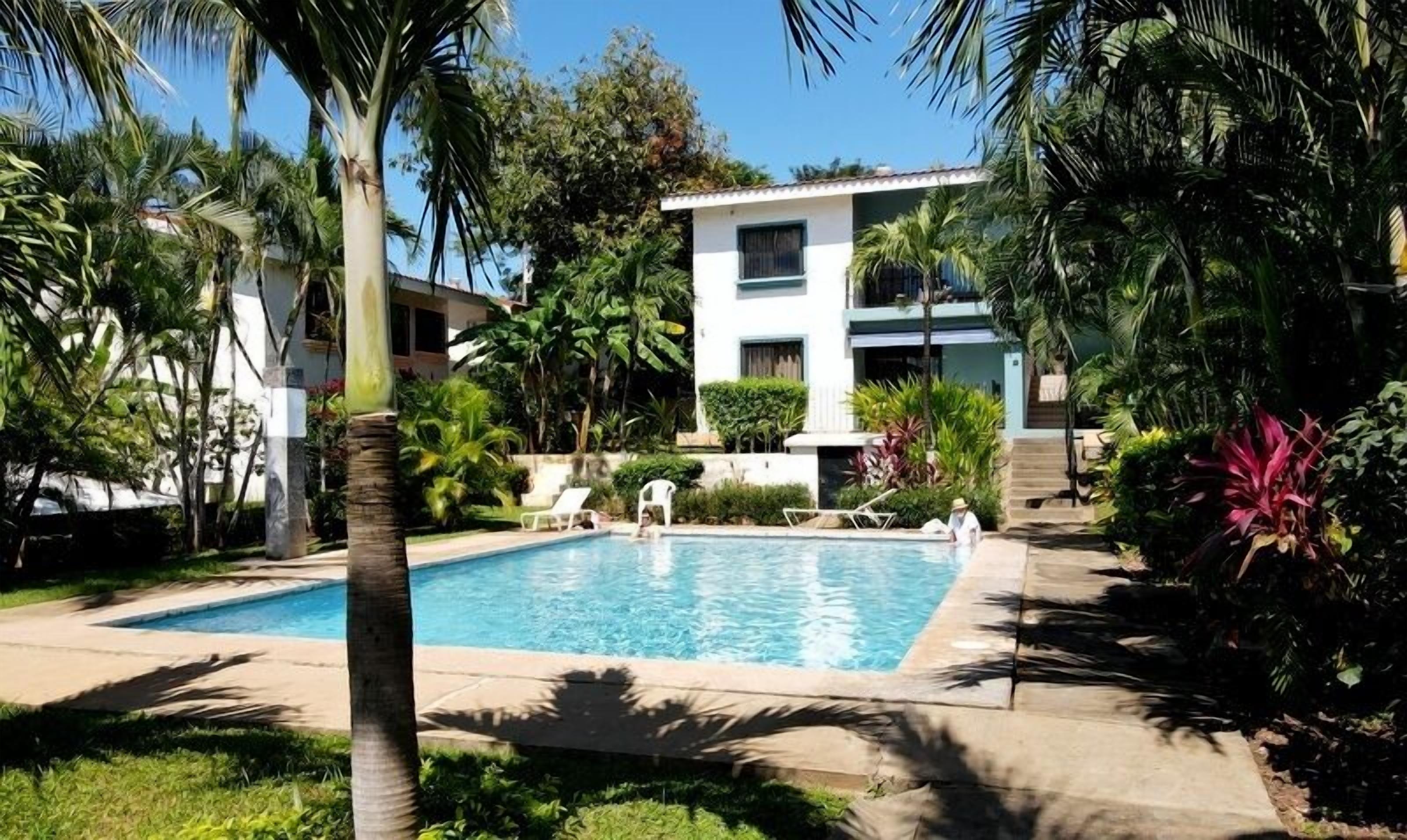 A swimming pool surrounded by lush greenery and a two-story building with a balcony.