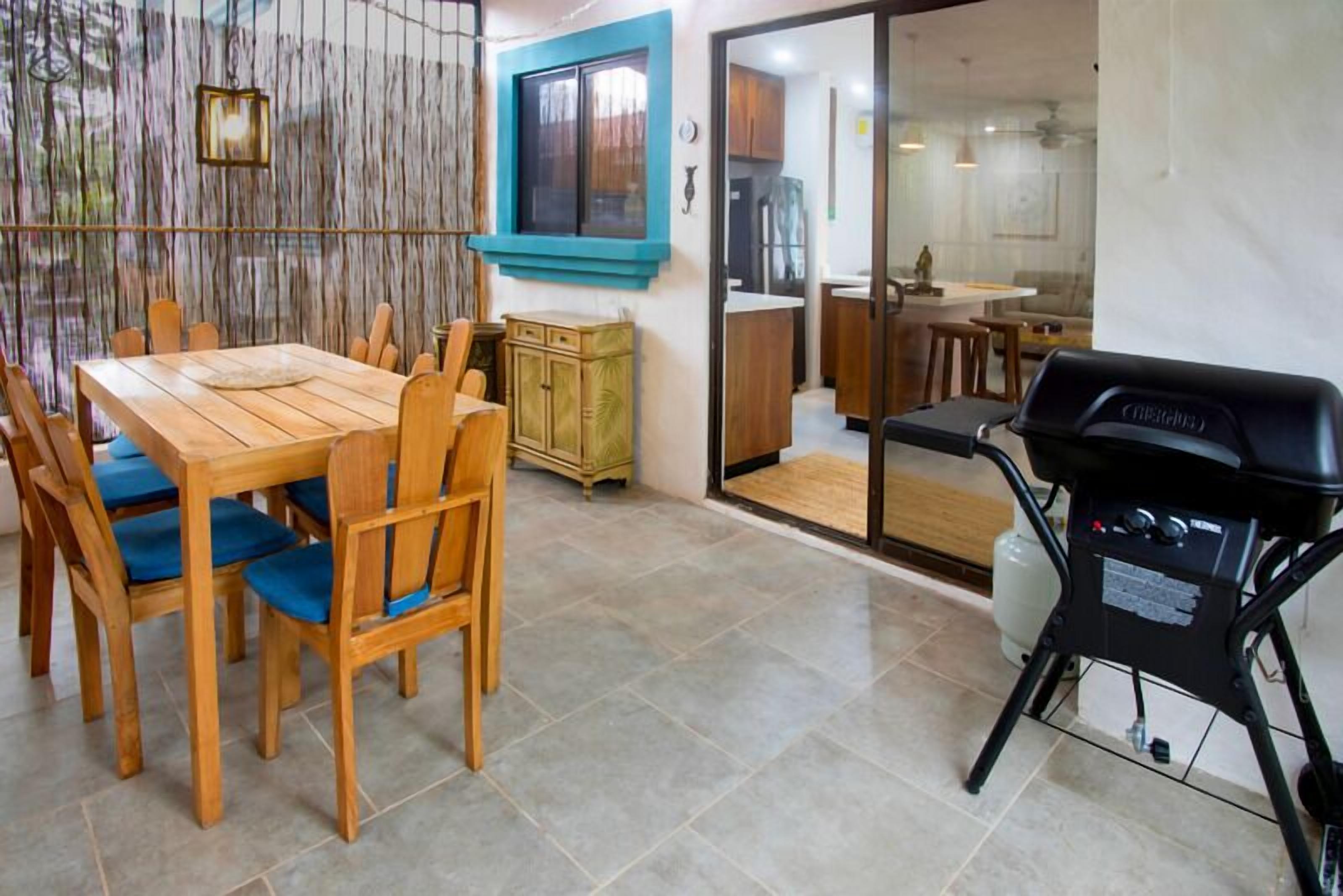 Un'area pranzo con tavolo e sedie in legno, una cucina visibile attraverso una porta in vetro e un barbecue all'esterno.