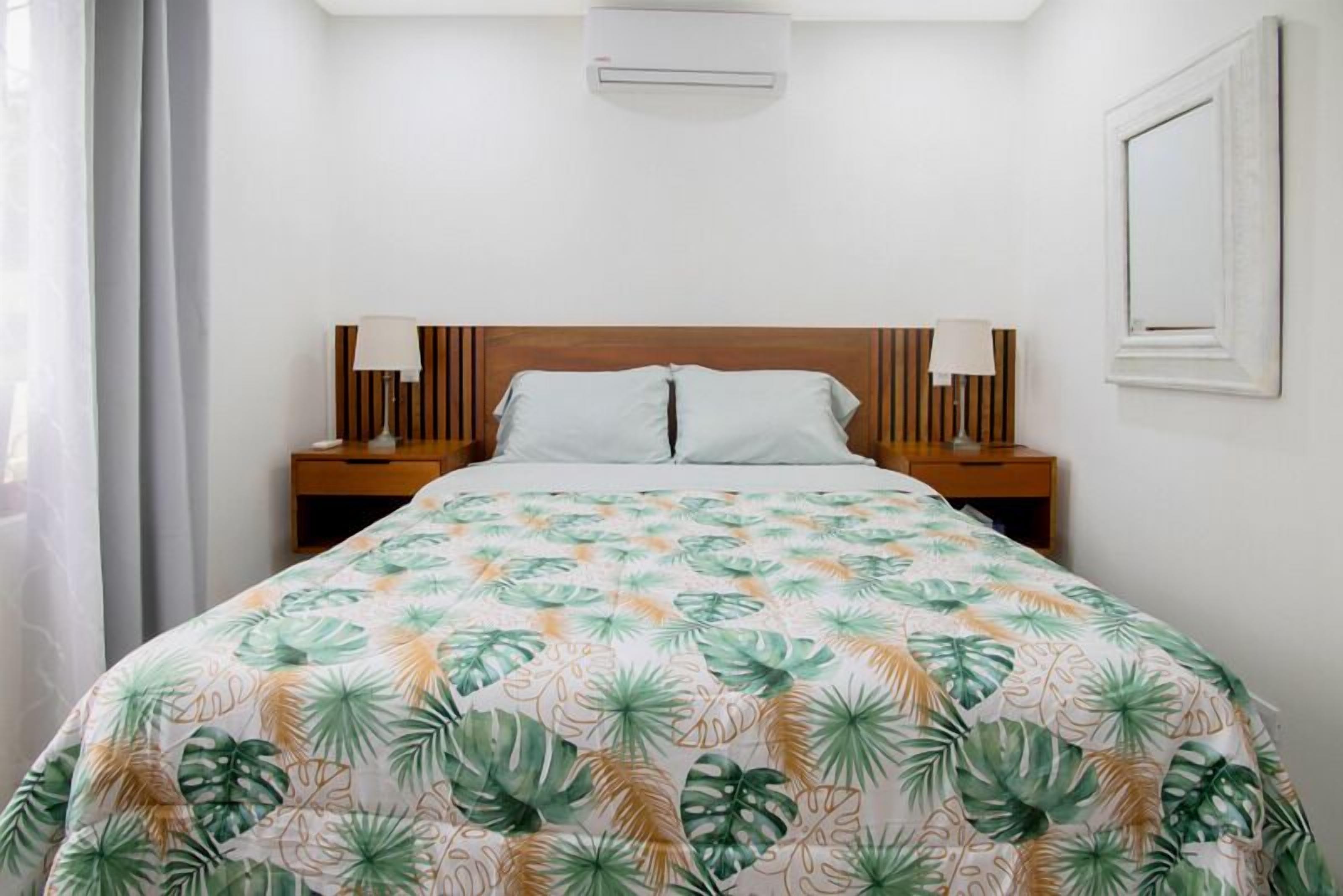 A neatly made bed with a tropical leaf pattern bedspread, a wooden headboard, and bedside lamps.