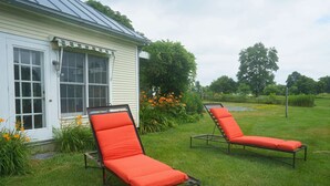Two outdoor lounge chairs with metal frames and orange cushions in front of a house with a white exterior and a green roof.