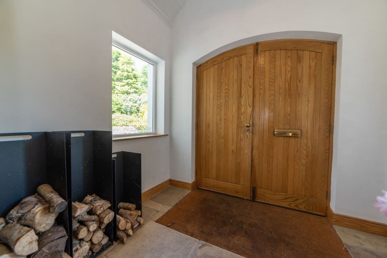 A room with a large wooden door, a window with a view of greenery, and a stack of firewood.