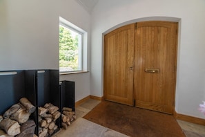 Una habitación con una puerta de madera grande, una ventana que da a zonas verdes, y una pila de leña.