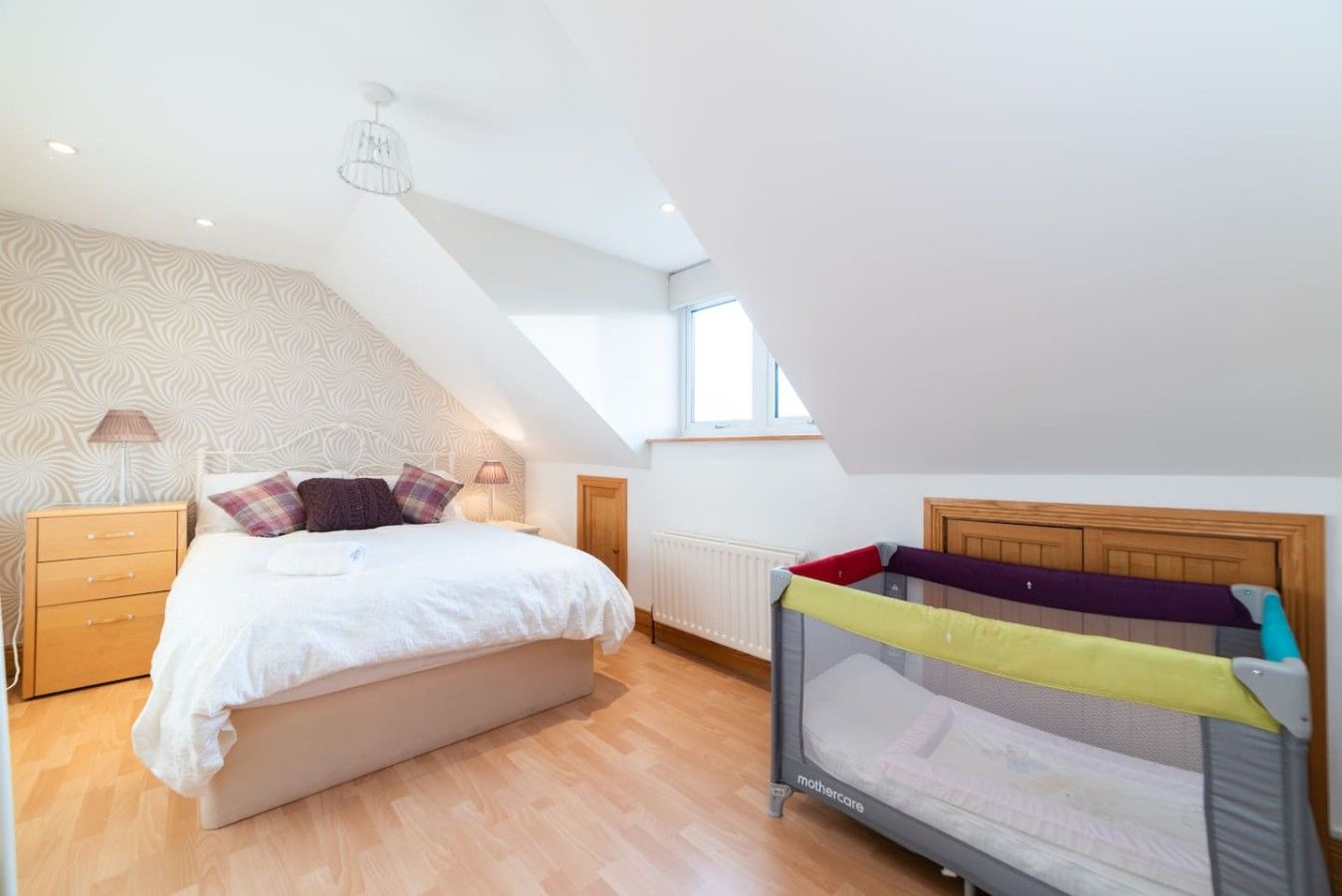 A bedroom with a sloped ceiling, a bed, a nightstand, a lamp, a window, and a crib.
