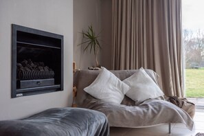 Una sala de estar con un sofá, una chimenea, una planta y vistas a través de una ventana con cortinas.