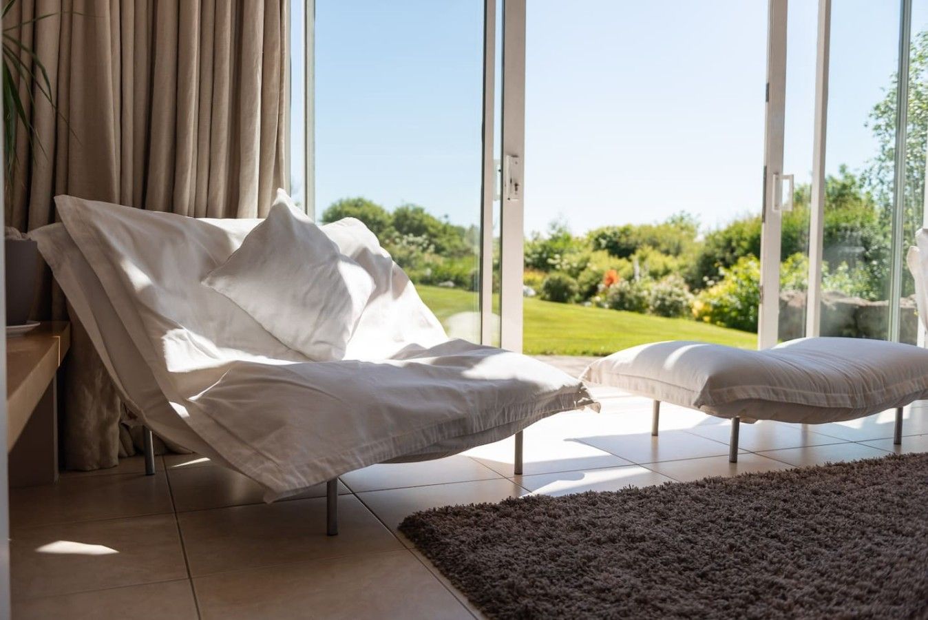 Ein modernes Schlafzimmer mit einem Bett, einem Nachttisch und Blick auf einen Garten durch große Fenster.