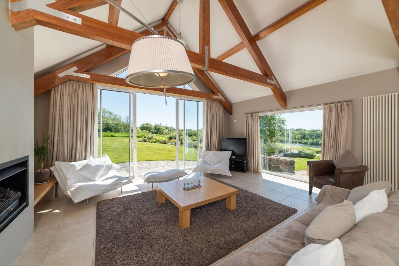 Ein geräumiges Wohnzimmer mit hoher Decke, Kamin, Fernseher und einem großen Fenster mit Blick auf eine grüne Landschaft.