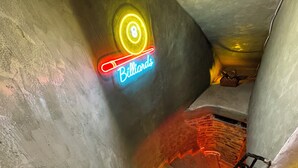 Un letrero de neón que dice "Billiards" en un estrecho rellano de escaleras tenuemente iluminado, con una pared texturizada y una barandilla de metal.