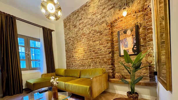 Una sala de estar con pared de ladrillo, un sofá amarillo, una mesa de centro de vidrio y un otoman marrón.