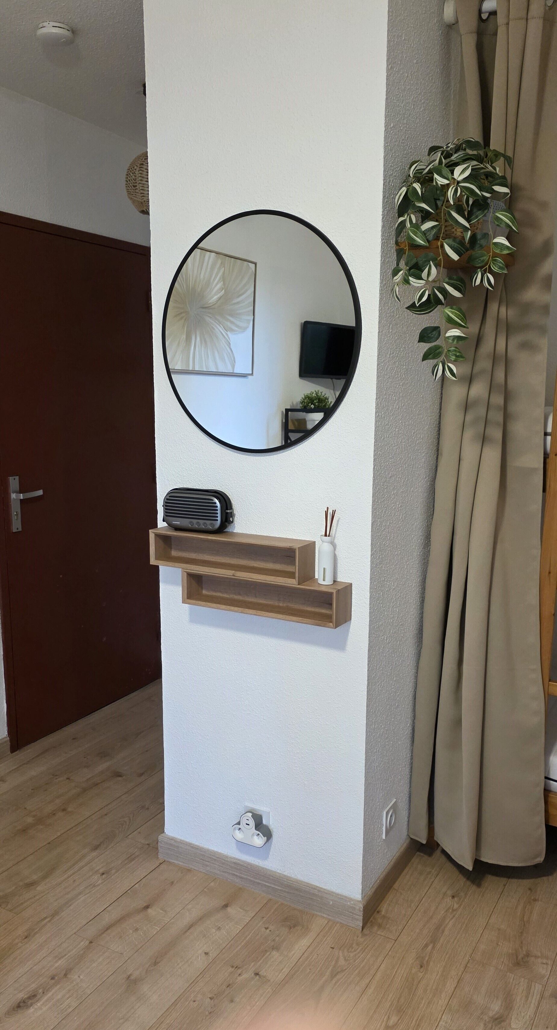 A room with a wooden floor, a wall-mounted shelf with a speaker and a small vase, a round mirror, and a potted plant.