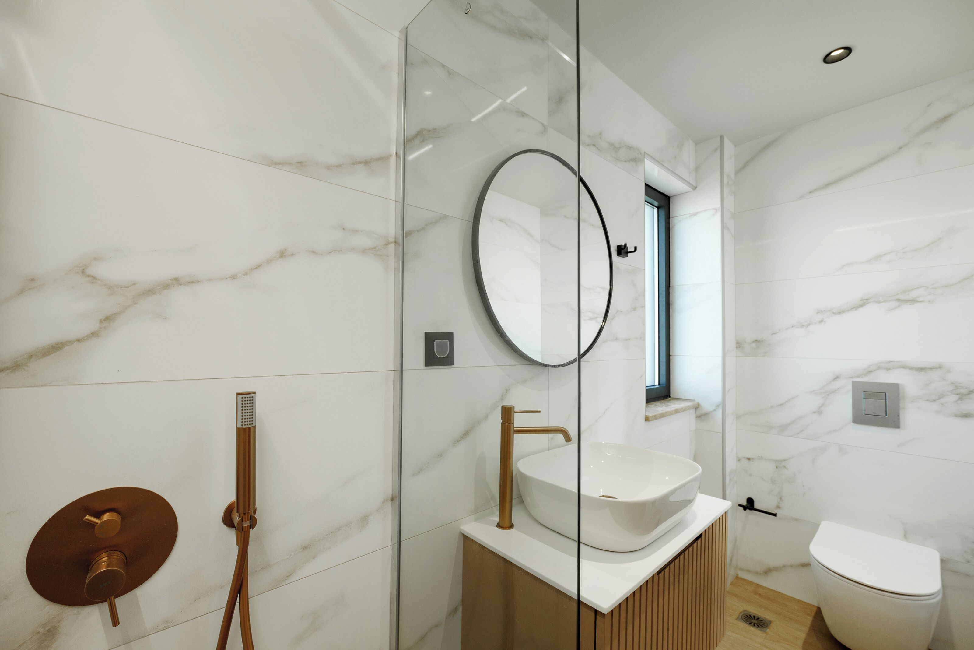 Une salle de bain moderne avec une douche à parois de verre, un miroir rond et un lavabo blanc avec un meuble-vasque en bois.