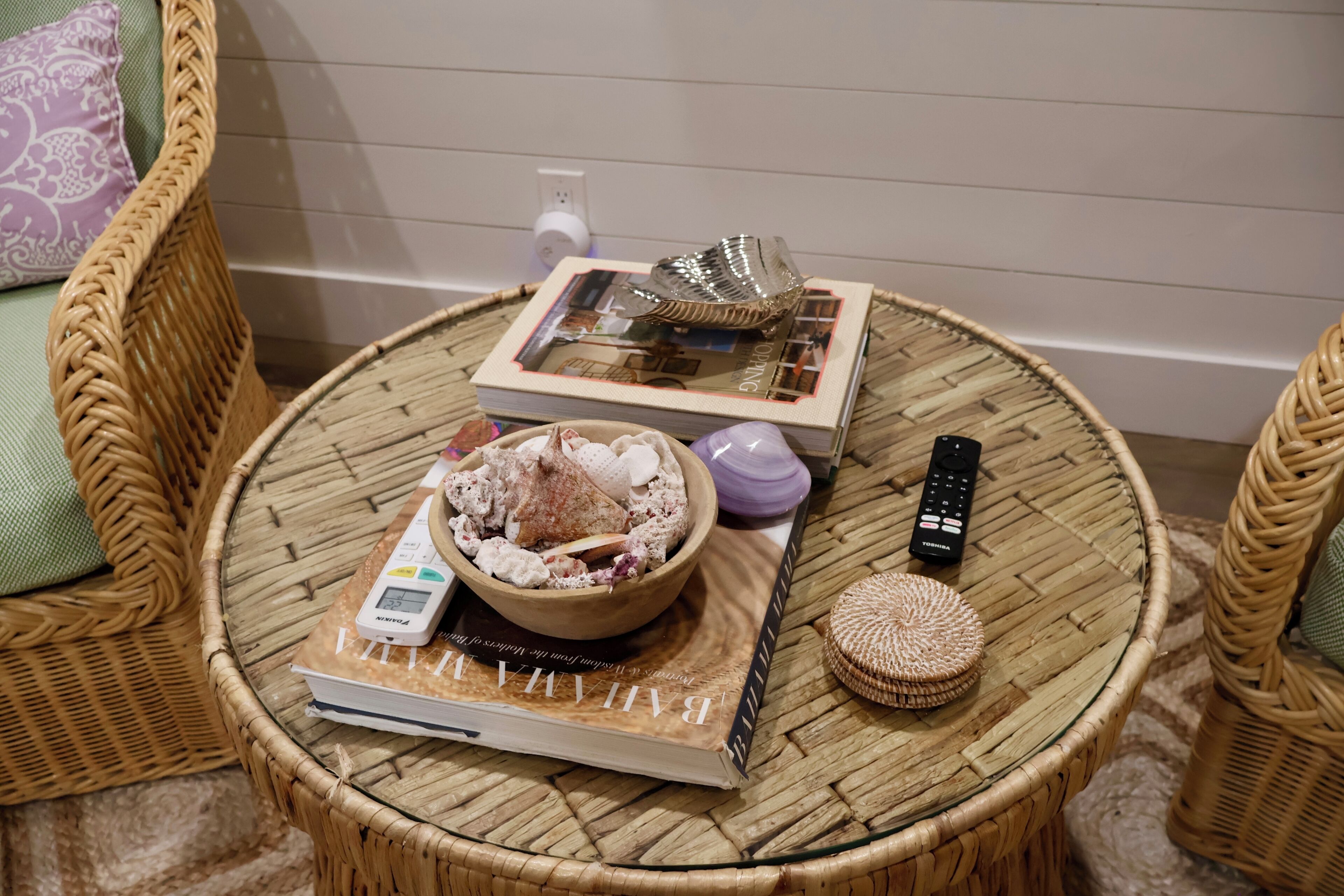 A round wicker table with a book, a bowl of snacks, a remote control, a small bottle, and a decorative item.