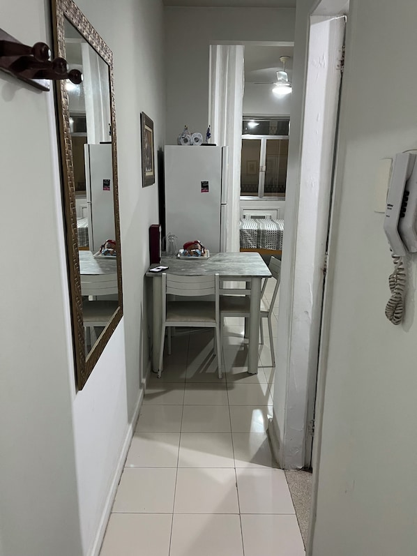 A narrow hallway with a white tiled floor, a white table with chairs, a refrigerator, and a telephone.