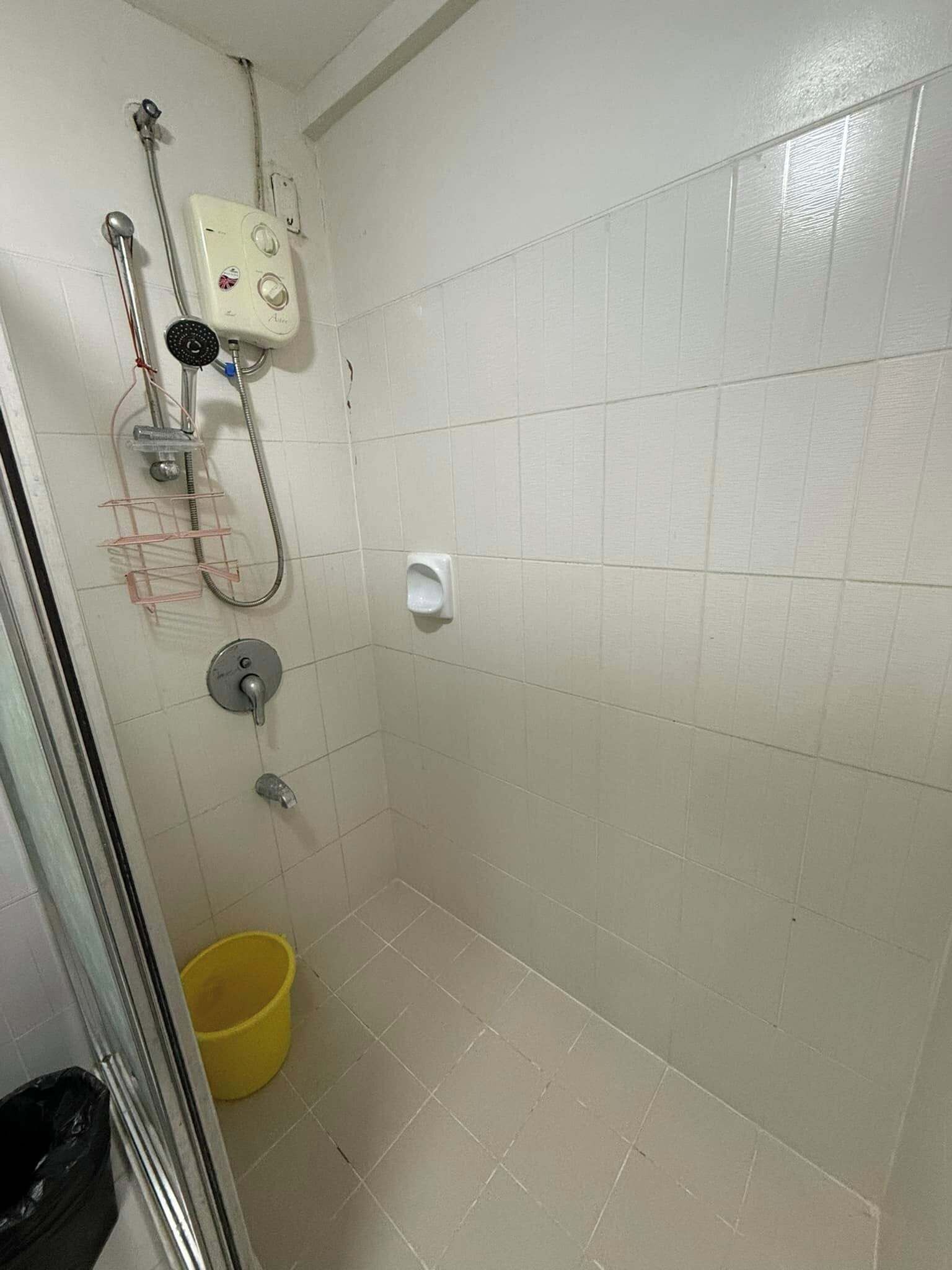 Ett litet, kaklat duschutrymme med ett handhållet duschmunstycke, en väggmonterad kontrollpanel och en gul hink på golvet.