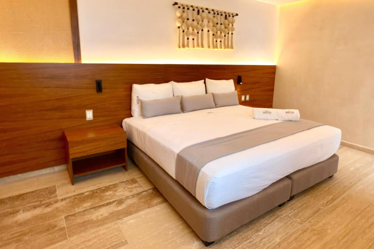 Una habitación de hotel moderna con cabecera de madera, una cama grande con edredón gris y blanco, una mesita de noche y una lámpara de pared.