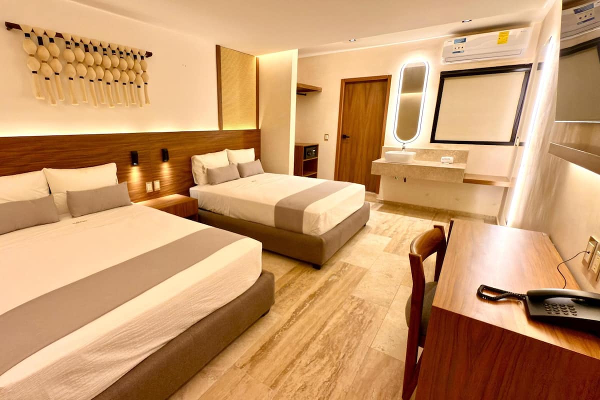 Habitación de hotel con dos camas, cabecero de madera, mesita de noche, escritorio con teléfono y baño con espejo y lavamanos.