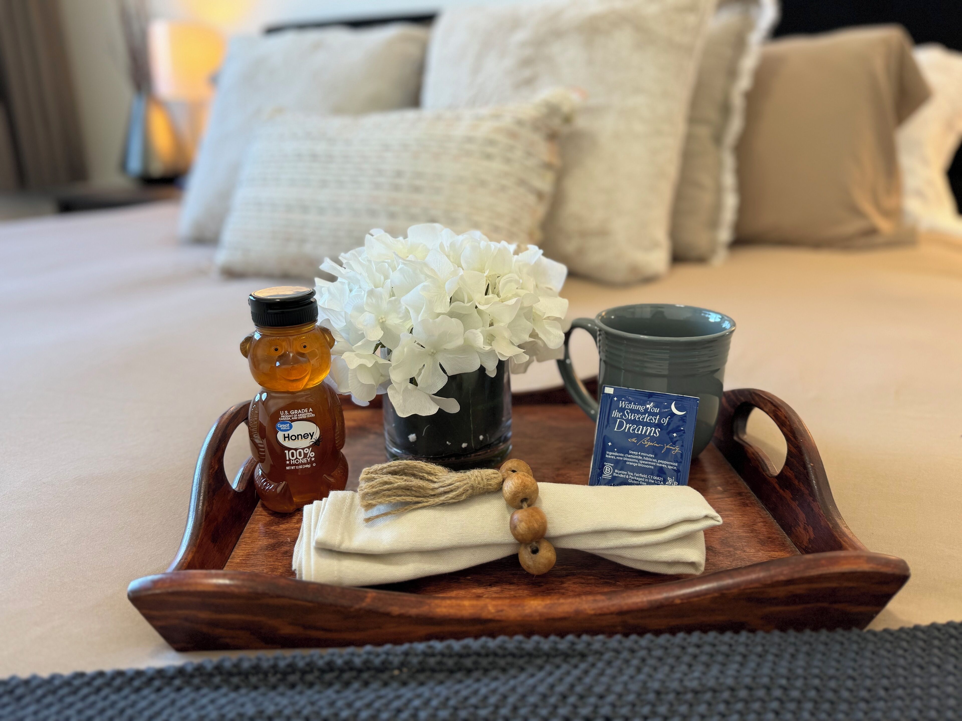 Un vassoio di legno con miele, una tazza, un biglietto e un vaso di fiori bianchi su un letto.