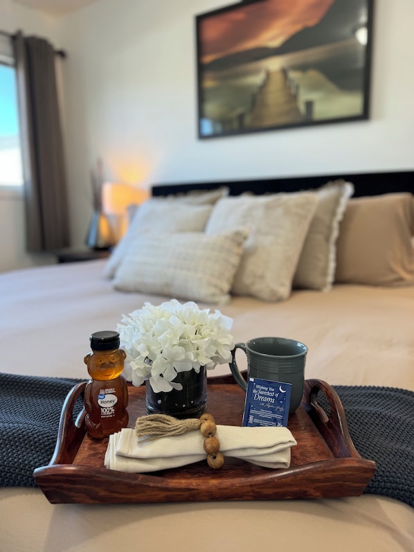 Una cama bien hecha con una bandeja decorativa al frente, que contiene un tarro de miel, un jarrón con flores blancas, una taza y una tarjeta.