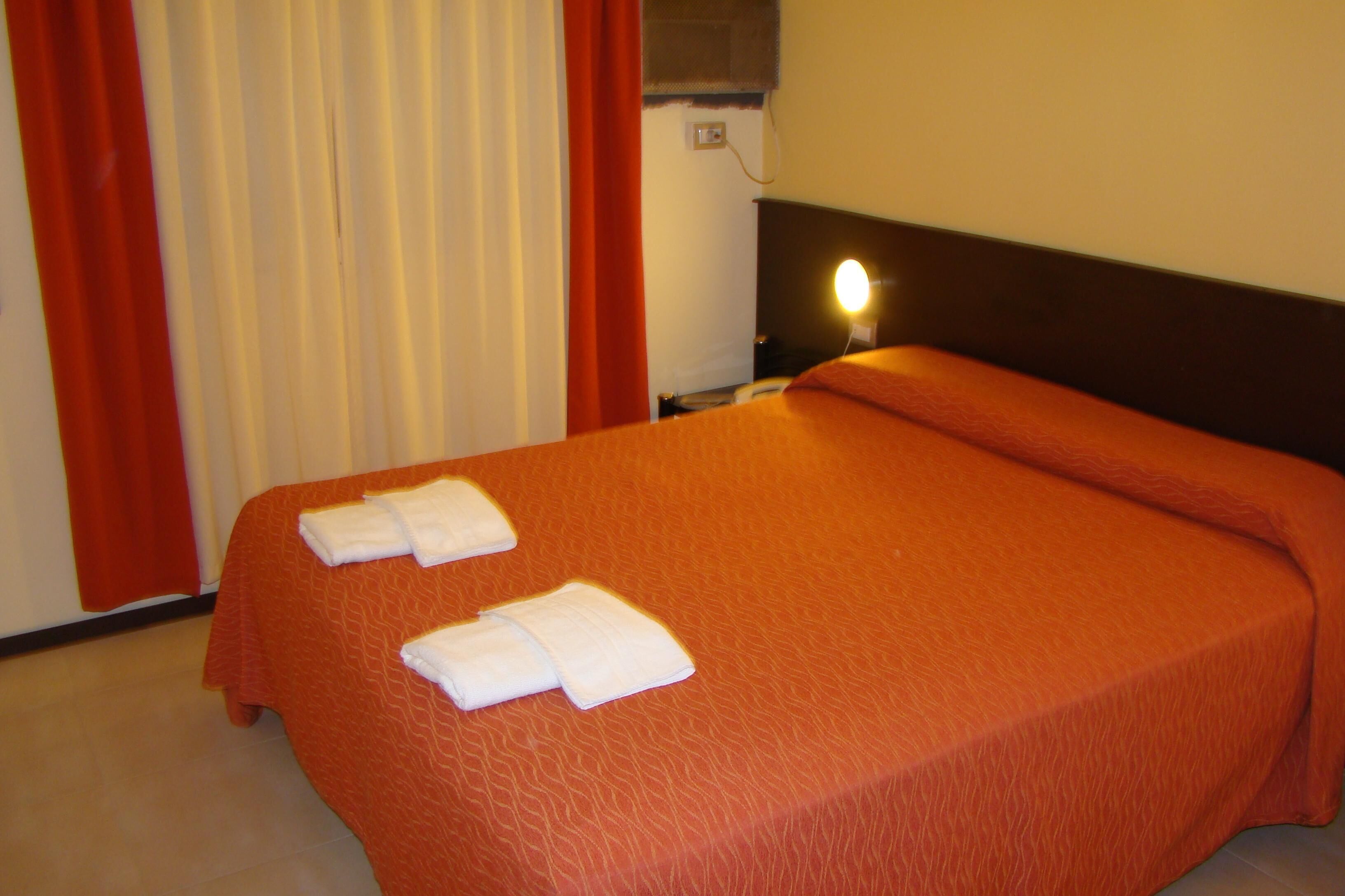 A hotel room with a bed, red curtains, a wall socket, and a bedside lamp.