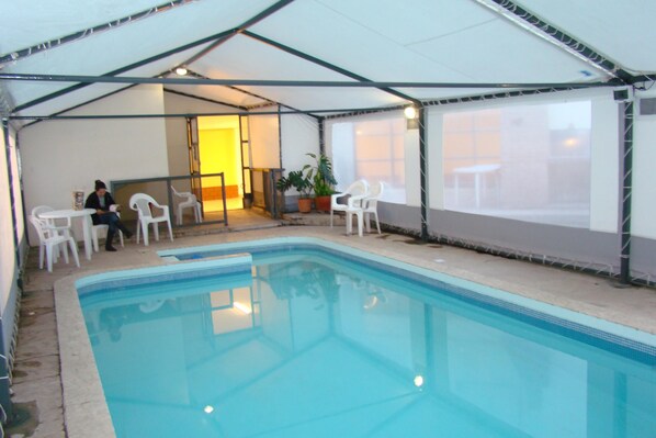 An indoor swimming pool with a covered area, white chairs, and a table.