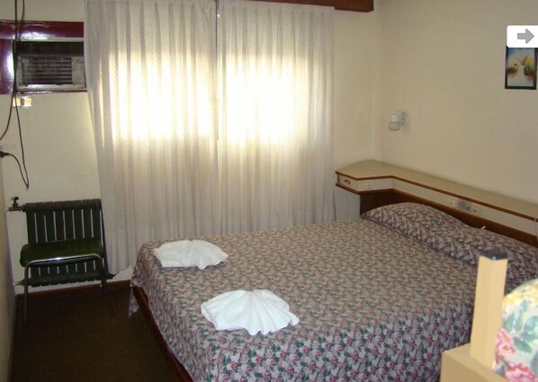 A hotel room with a bed, a nightstand, a radiator, and a window with curtains.