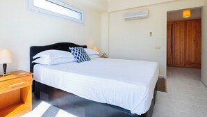 Een slaapkamer met een bed, nachtkastje, lamp, raam, airconditioning en een houten garderobe.