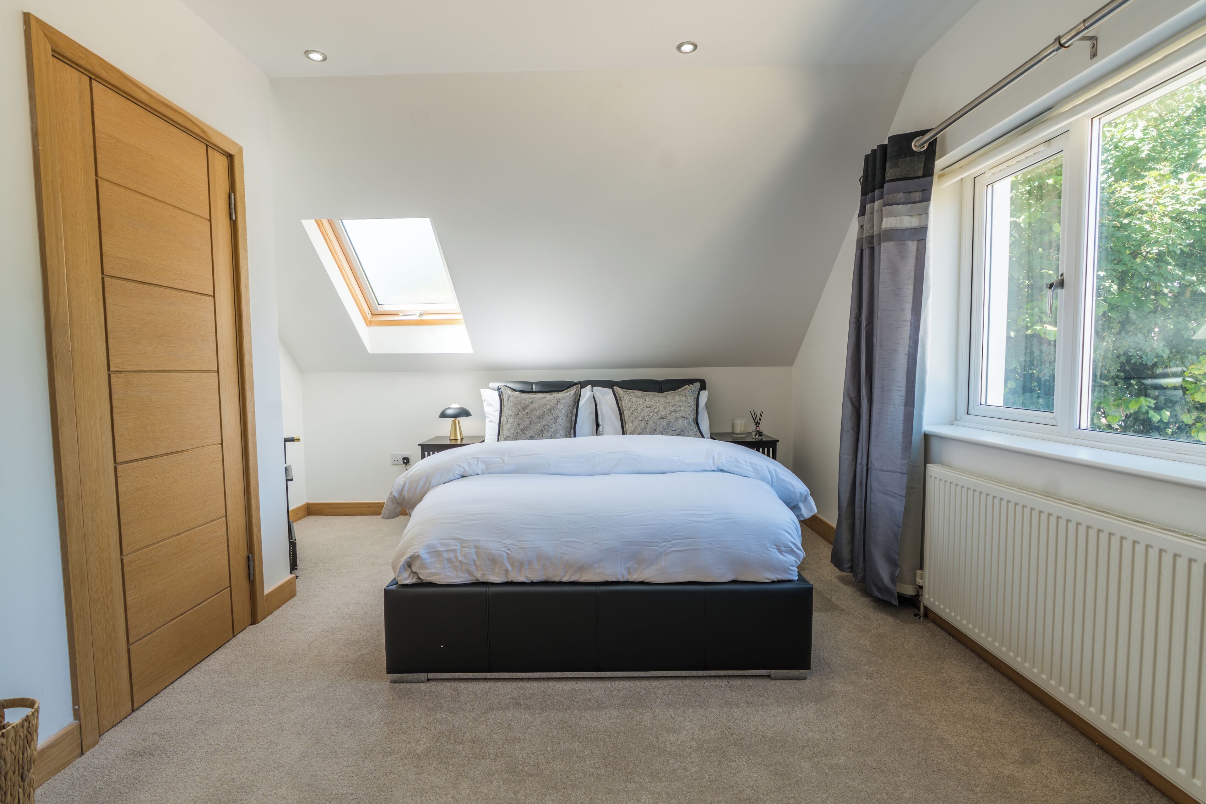 A bedroom with a skylight, a large bed, a window with curtains, and a radiator.