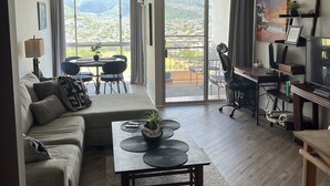 Un vaste salon doté d’une grande fenêtre offrant une vue sur les montagnes et le paysage urbain. On y trouve un canapé, une table basse avec une plante et un bureau avec un ordinateur.
