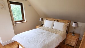 A bedroom with a wooden floor, a bed with white bedding, two bedside tables with lamps, a window with curtains, and a ceiling light fixture.