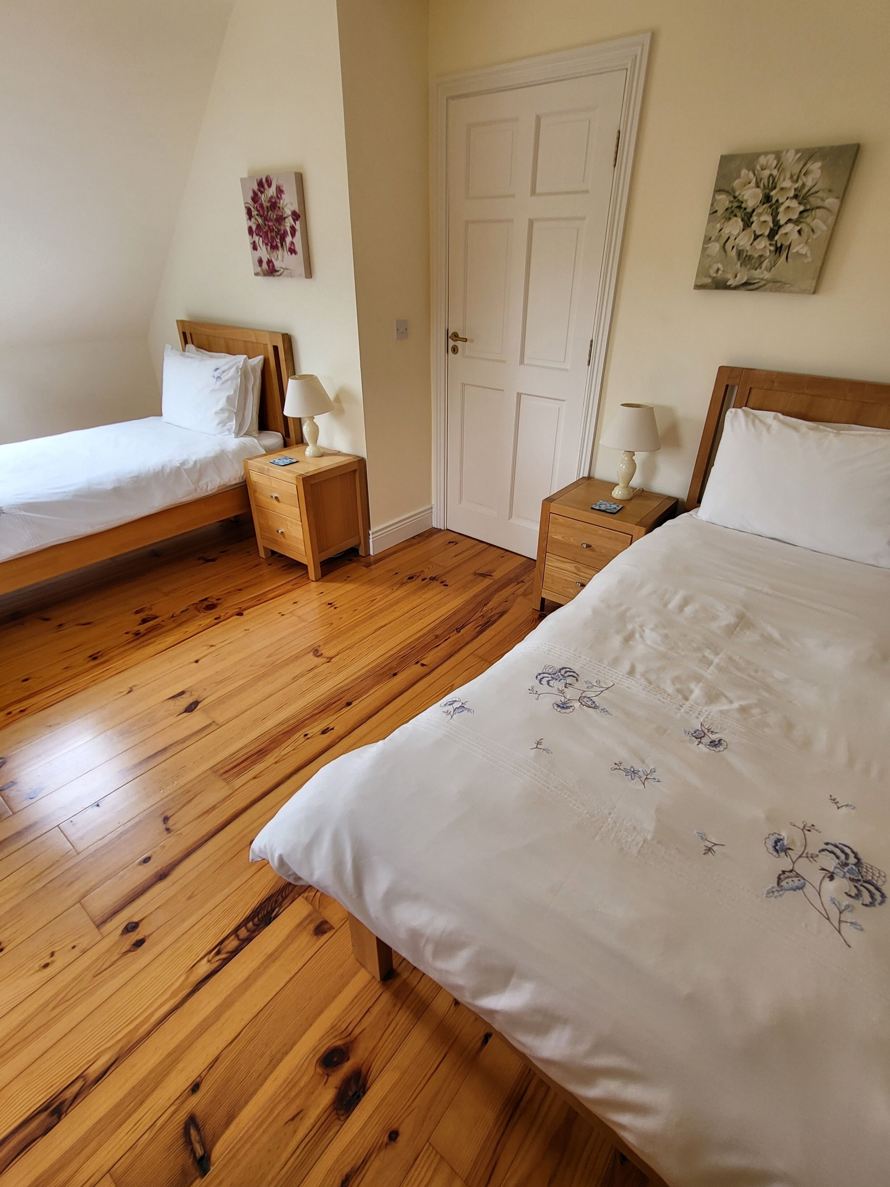 Een slaapkamer met twee eenpersoonsbedden, houten vloeren, een witte deur en bloemistische kunst aan de muren.
