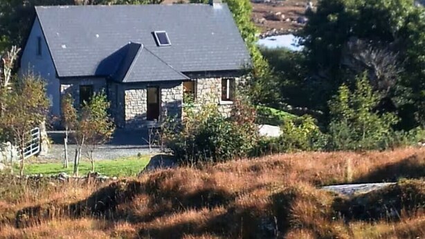 A stone house with a dark roof, surrounded by a grassy landscape and trees.