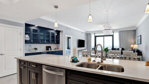A modern kitchen with dark cabinets, a large island with a sink, pendant lighting, and a dining area with a TV and seating.