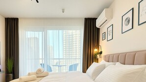 A hotel room with a large bed, a bedside table with a lamp, a window with curtains, and a view of buildings outside.