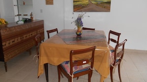 Un coin repas avec une table recouverte d’une nappe jaune, entourée de chaises en bois. Un vase rempli de fleurs est posé sur la table.