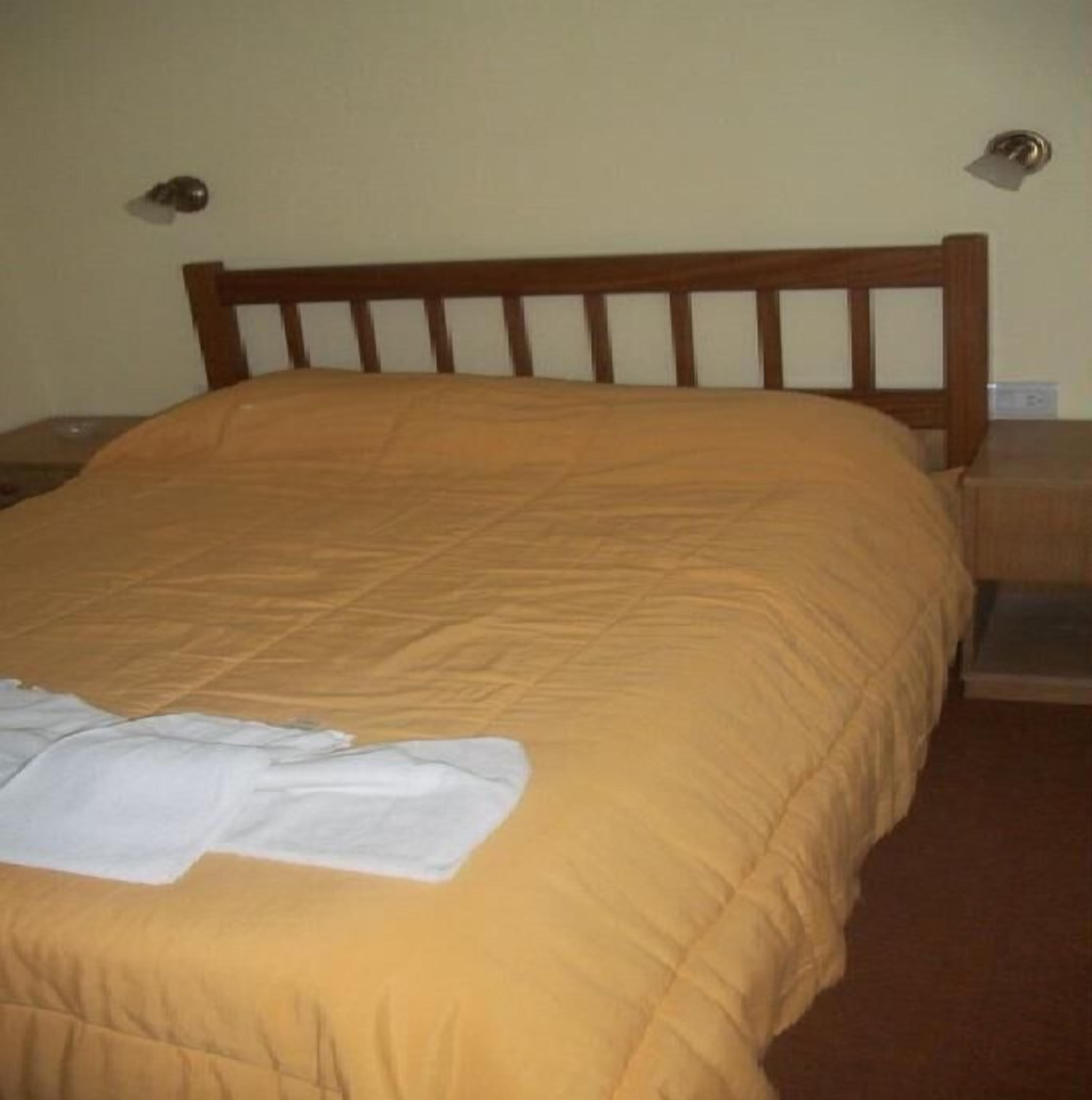 A bed with a wooden headboard, a white blanket, and a brown carpeted floor.