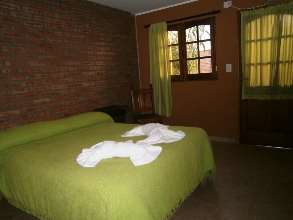 A room with a brick wall, a bed with a green bedspread, a window with yellow curtains, and a wooden chair.